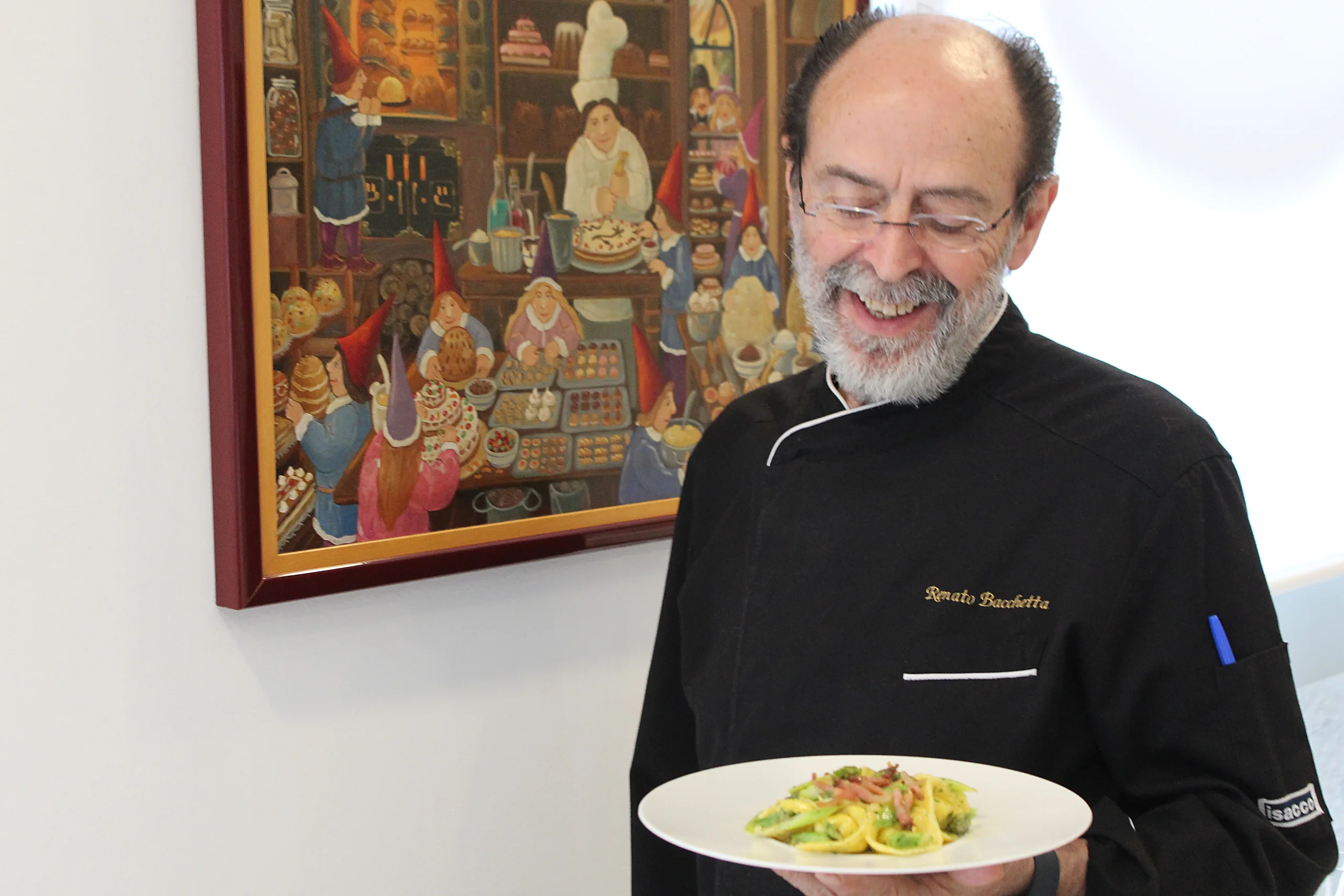 Lo Chef Renato Bacchetta sorride con un piatto in mano accanto a un quadro della sala